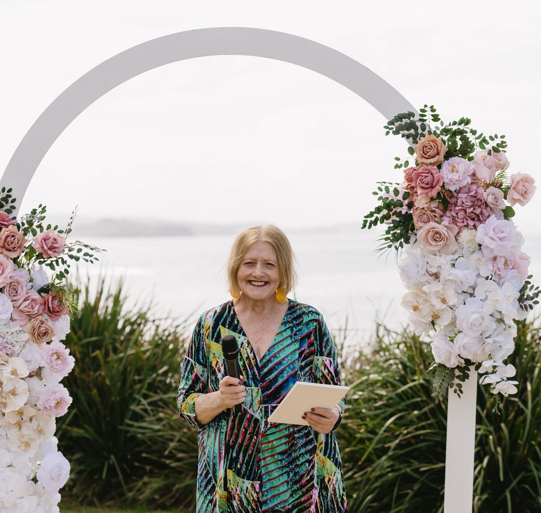 Kathy stands beneath a floral arch by the beach, holding a microphone and notepad outdoors.