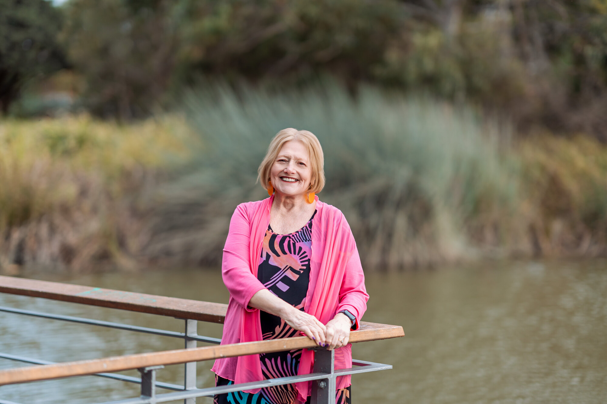 Kathy Rozmeta Celebrant, older woman in pink cardigan, smiles by pond with greenery in the background.